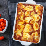A delicious-looking Eggplant Parmesan, fresh from the oven in a ceramic baking dish. The eggplant is golden brown and crispy, and the cheese is melted and gooey. The eggplant is sitting next to a potential garnish of cherry tomoes