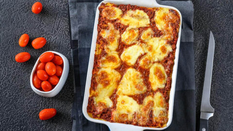 A delicious-looking Eggplant Parmesan, fresh from the oven in a ceramic baking dish. The eggplant is golden brown and crispy, and the cheese is melted and gooey. The eggplant is sitting next to a potential garnish of cherry tomoes
