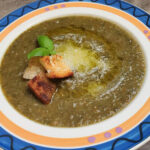A bowl of vibrant veggie puree soup garnished with crispy bread, drizzles of olive oil, and parmesan shavings, representing an Authentic Italian Recipes dish.